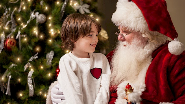 A child sits on Santa's lap next to a decorated Christmas tree, both smiling warmly, with festive lights glowing.