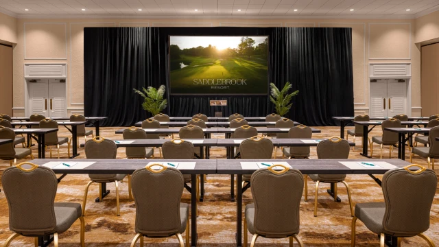 A conference room set up with rows of tables and chairs facing a screen displaying