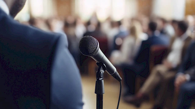A person stands near a microphone facing an audience in a blurred background, suggesting a presentation or public speaking event.
