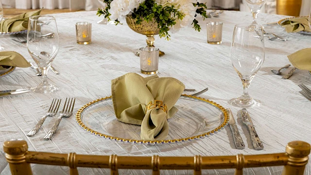 A table elegantly set with a gold-rimmed plate, folded napkin, silverware, wine glasses, and floral centerpiece on a white tablecloth.