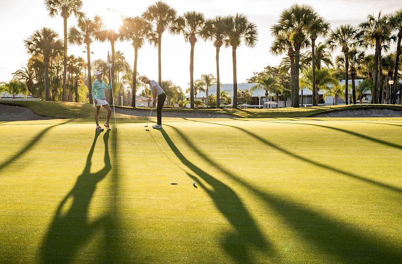 Two people are on a golf course with palm trees at sunset, casting long shadows